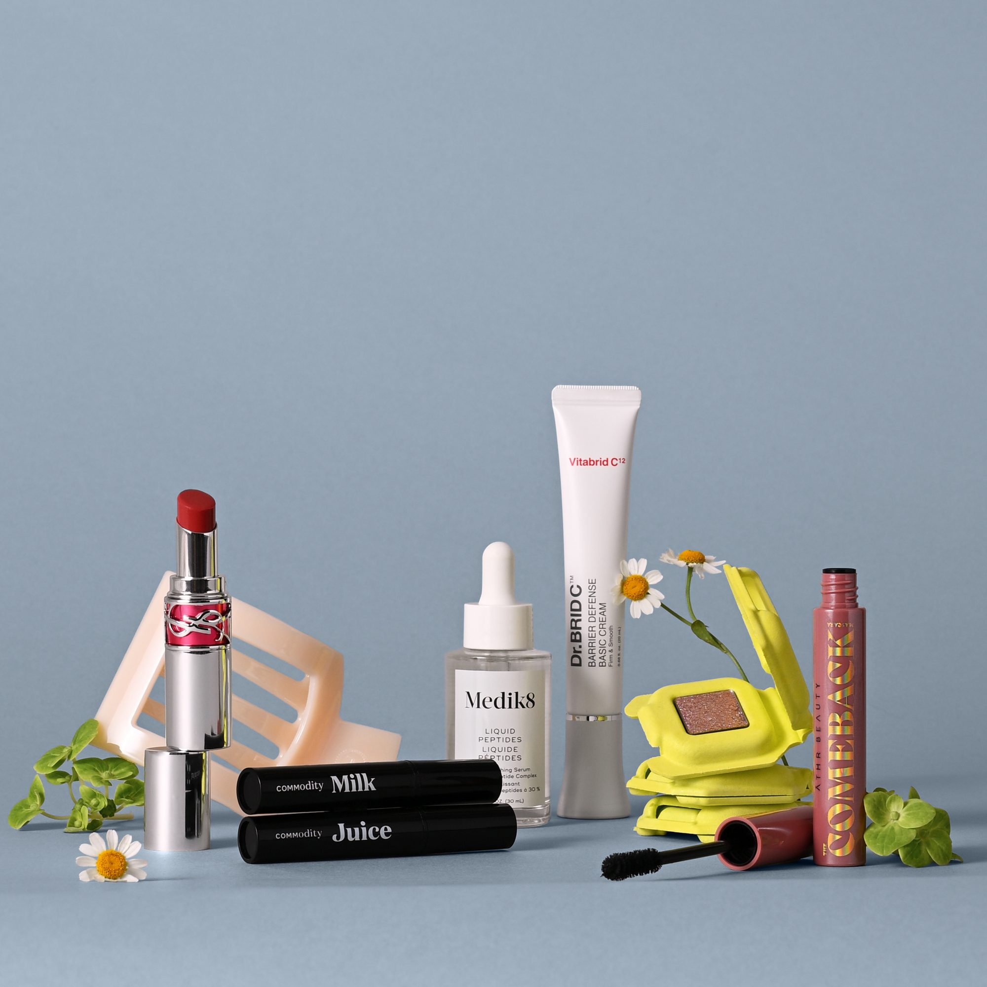 Lineup of beauty products including red lipstick, mascaras, white tube, serum bottle, eyeshadow palette, and spray with flowers and greenery on blue surface