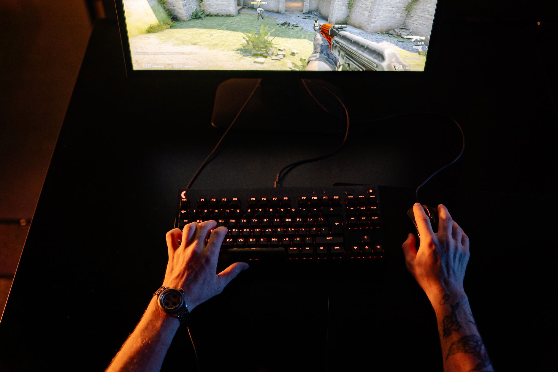 Person's hands on illuminated gaming keyboard with first-person shooter game displayed on monitor above