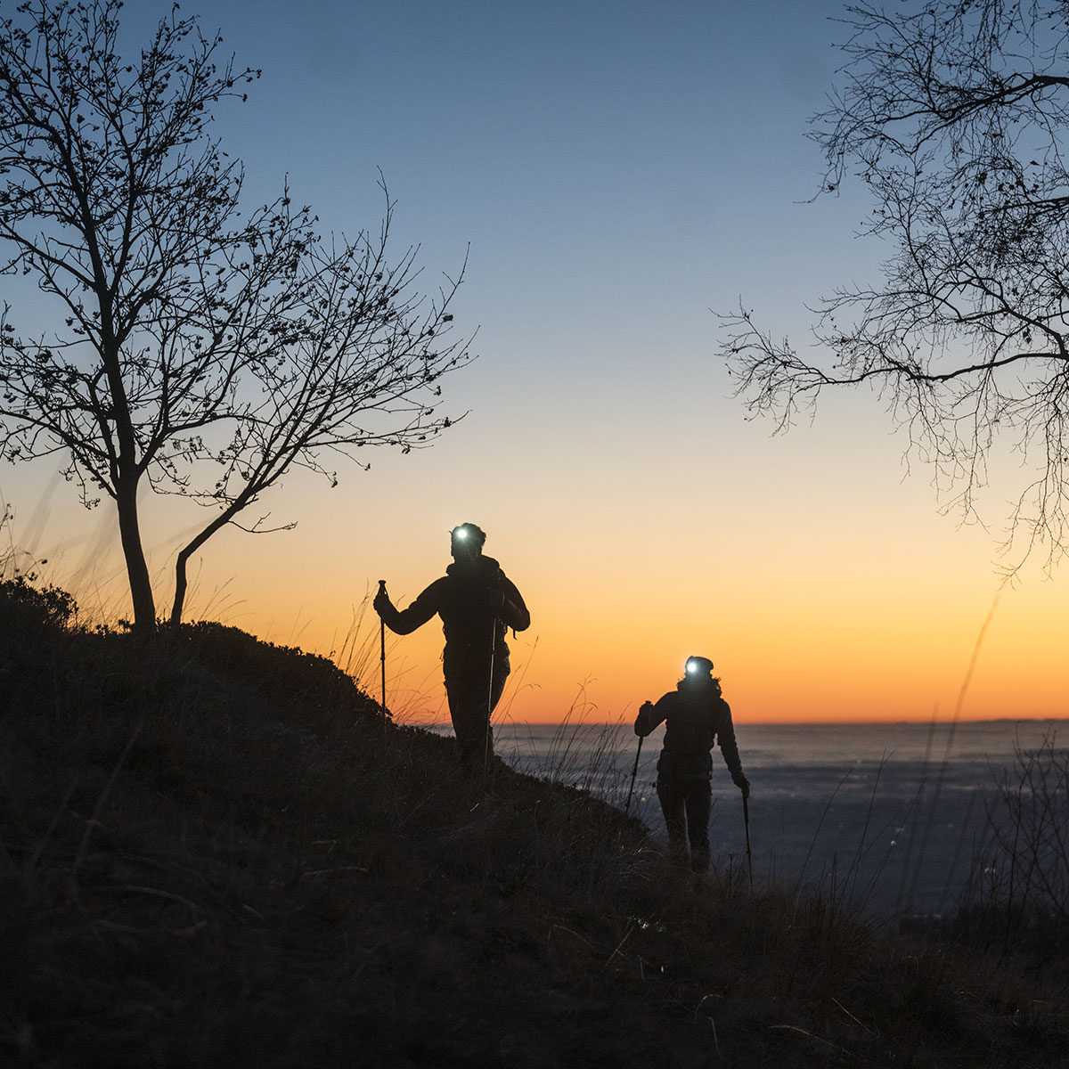 Due escursionisti con torce frontali camminano su un pendio al tramonto vicino al mare.