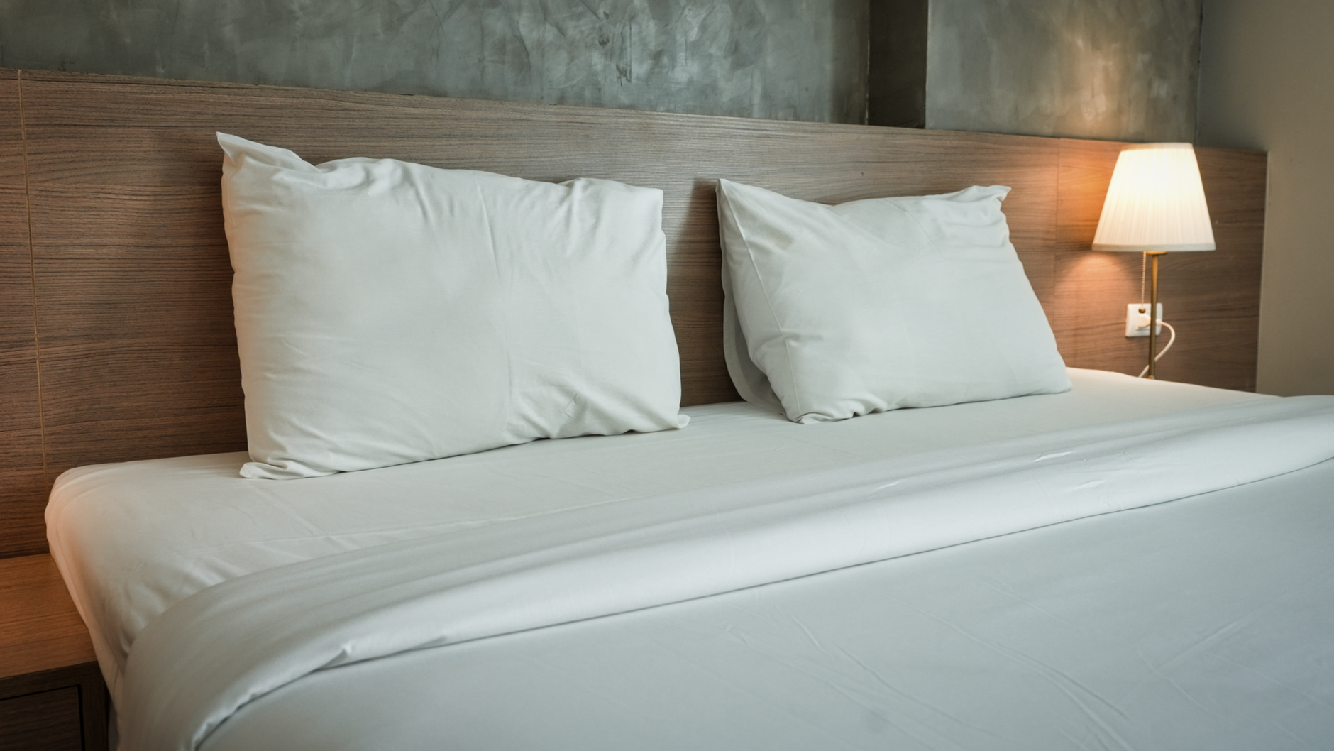 Letto matrimoniale con testata in legno, due cuscini bianchi, lampada da comodino e parete in cemento