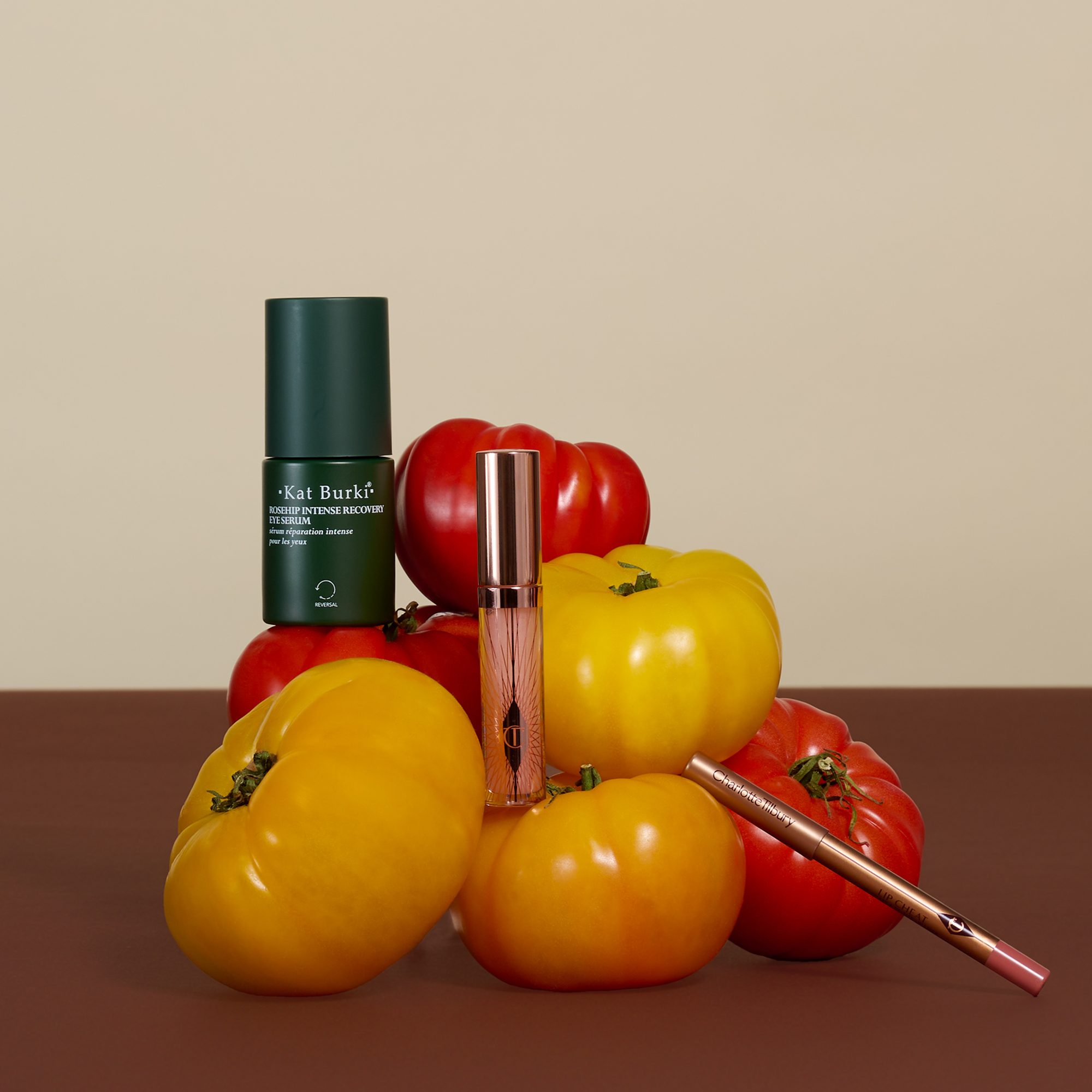 Green serum bottle and pink lip gloss displayed with red and yellow tomatoes and a makeup brush on brown surface