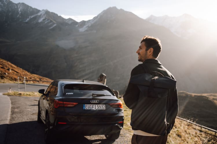 Mann in schwarzer Jacke steht neben dunkler Limousine in Berglandschaft mit Seenblick
