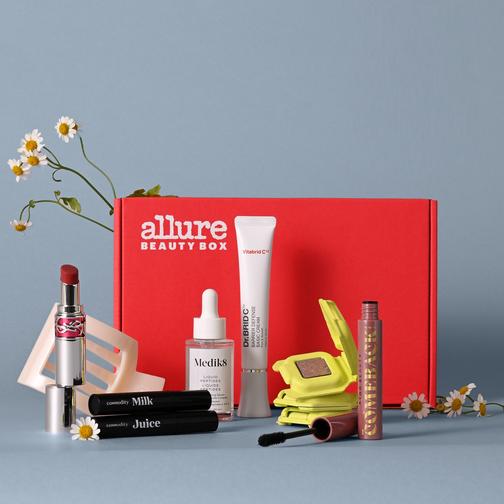 Red subscription box with beauty products including red lipstick, mascaras, serum, eyeshadow palette, and spray with daisies on blue background